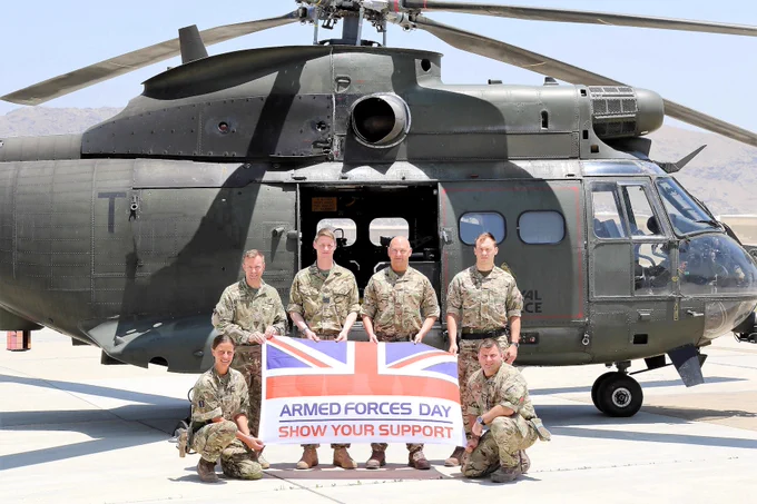 Armed Forces personnel unfurl an Armed Forces Day flag in front of a helicopter.