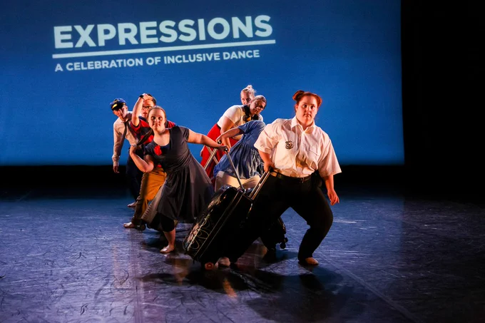 Ability performing at Expressions 2019. A group of dancers are stood in a line on stage looking out at the audience. Behind them is a blue backdrop with the title 'Expressions: a celebration of inclusive dance'. Photo by Brian Slater.