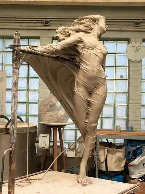 Photograph of art studio setting featuring a large sculpture of a female standing figure as if in walking movement with one leg raised and arms outstretched, the figure appears naked under a soft veil like material which forms a cloak behind her, the figure has long wavey hair as if blown by the wind, she faces forwards and looks right, the sculpture is placed on wooden plinth, behind the sculpture are glass panels in window frames and below are various equipaments, bags, tables and wires in the studio