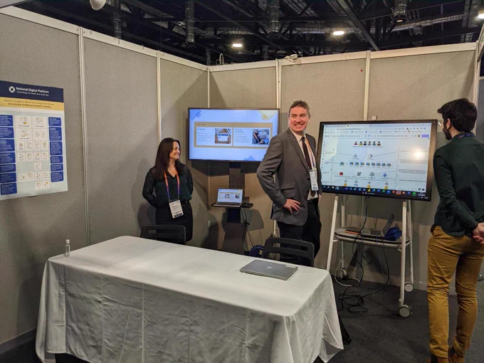 National Digital Platform (NDP) team at their stand at the NHS Scotland conference in Aberdeen. In the photo there is a poster of NDP services, a rolling presentation on a screen and an interactive whiteboard. Three people laughing and smiling as the photo is taken.