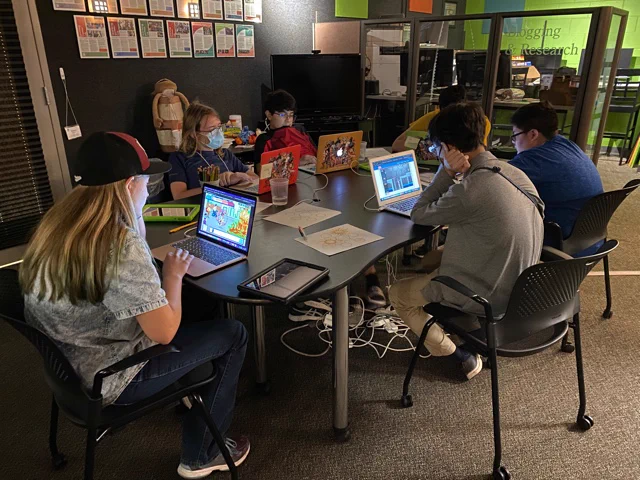 High school aged youth sit around a table playing learning games on laptops.