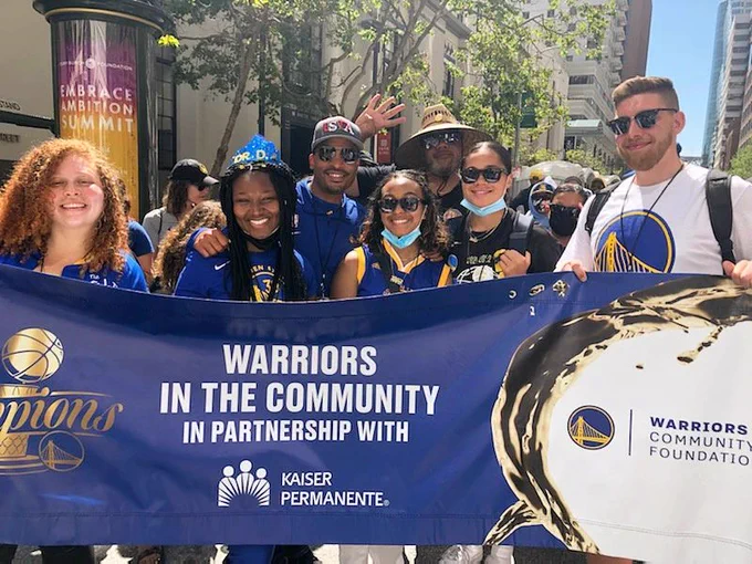 Smiling YMCA staff in Warriors t-shirts holding a banner that says "Warriors in the Community"