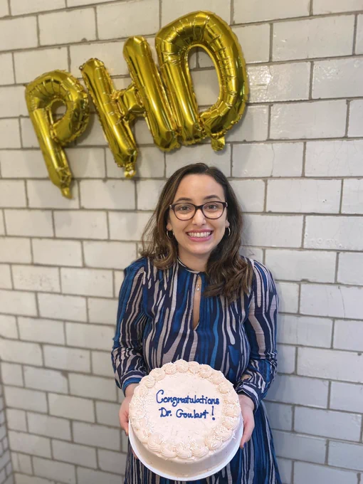 Inside picture, girl wearing a blue dress (me) holding a cake that reads “congrats Dr. Goulart” against a white brick wall with balloons of the letters PhD.