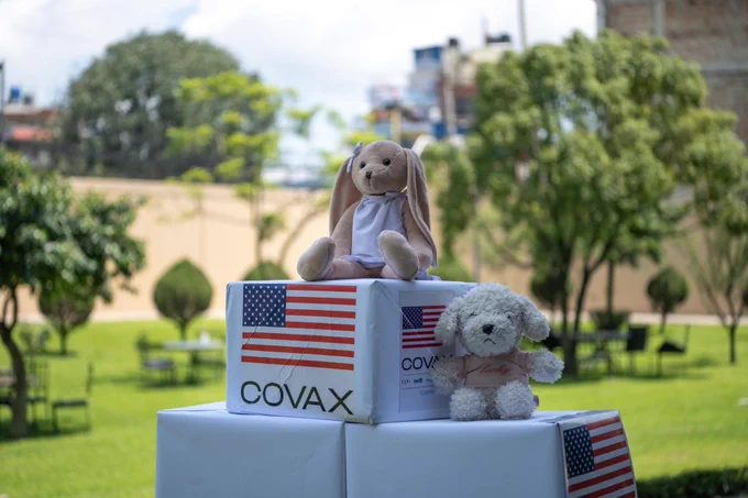 Stacked boxes are labeled with the American flag and the word COVAX, with two stuffed toys displayed on top.