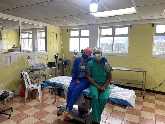 Two people sit on operating room table with masks, scrub hats, and goggles on their heads. 