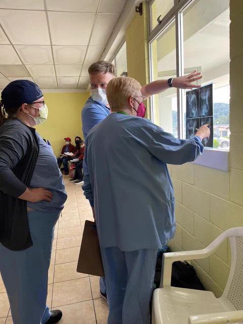Three physicians in blue scrubs review x-rays held to window light. 