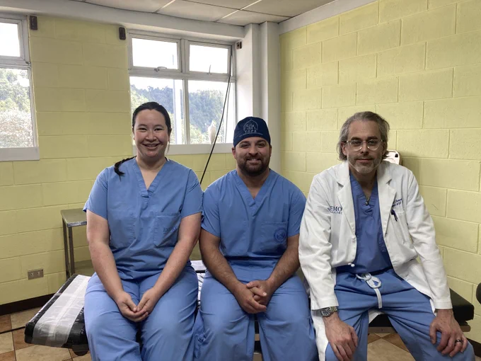 Two faculty and one resident sit smiling on an operating room table. 