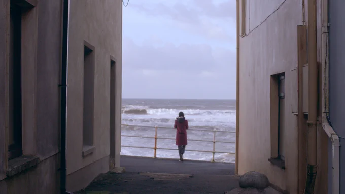 Image of person leaning on a railing looking at waves in the sea. In the foreground are stone buildings. 