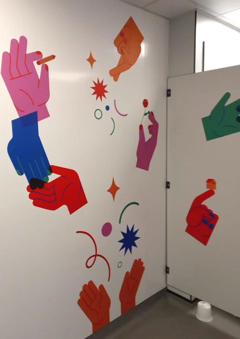 Vinyls of hands in different colours - in orange, red, pink, green and blue - are displayed on a white bathroom wall and door. One hand balances a little house, another one holds a pen and another one holds a flower.