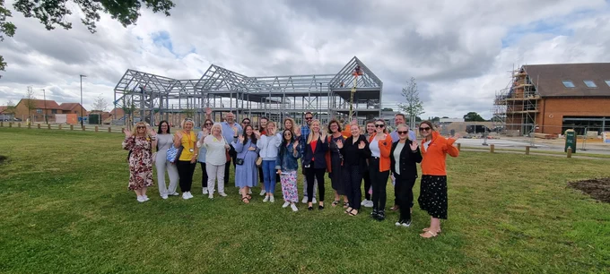 Group of 21 Fundraising Team members all waving and smiling to the camera in front of a building steel frame.