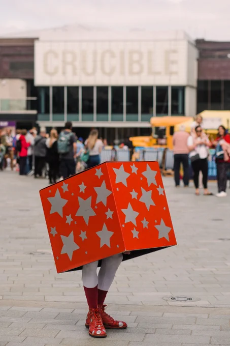 A red gift box decorated with stars stands on a pair of legs outside, in the background is a yellow bus and people in front of the Crucible Theatre.