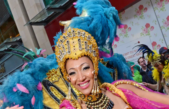 The image depicts the face of a smiling woman wearing a carnival costume with blue feathers, pink fabric and a golden headpiece.