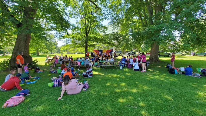 A crowd of parents, kids, and day camps on picnic tables and blankets on a lush, green lawn watching the Calithumpians perform a song in front of a colorful Red, Blue, and Yellow backdrop.
