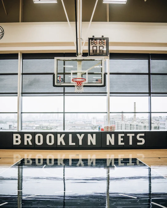 Photo of the Brooklyn Nets basketball practice court, looking at the basket from the free throw line. The wordmark Brooklyn Nets is visible in the background and the windows look out to Lower Manhattan