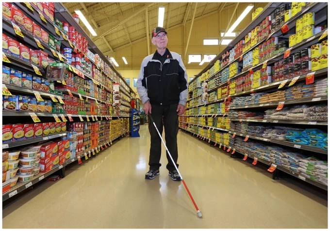 A man stands in a grocery store aisle with a white cane. 