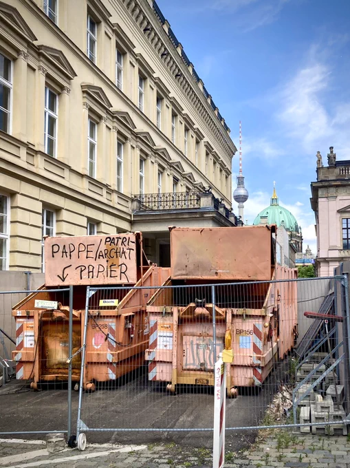 Hinter einem Bauzaun in Berlin Mitte steht ein Recycling Container. Darüber eine Tafel mit der Aufschrift Pappe, Patriarchat, Papier.