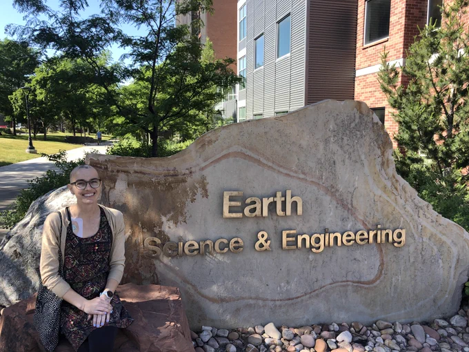 Photo of me wearing a black and floral print dress and bald head in front of a stone sign that reads "Earth Science & Engineering"