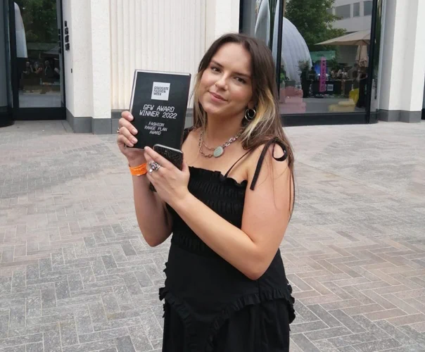 Fashion student Francesca Merrick outside a building during Graduate Fashion Week. She is holding a glass award which says "GFW Award Winner 2022" and "Fashion Range Plan Award"