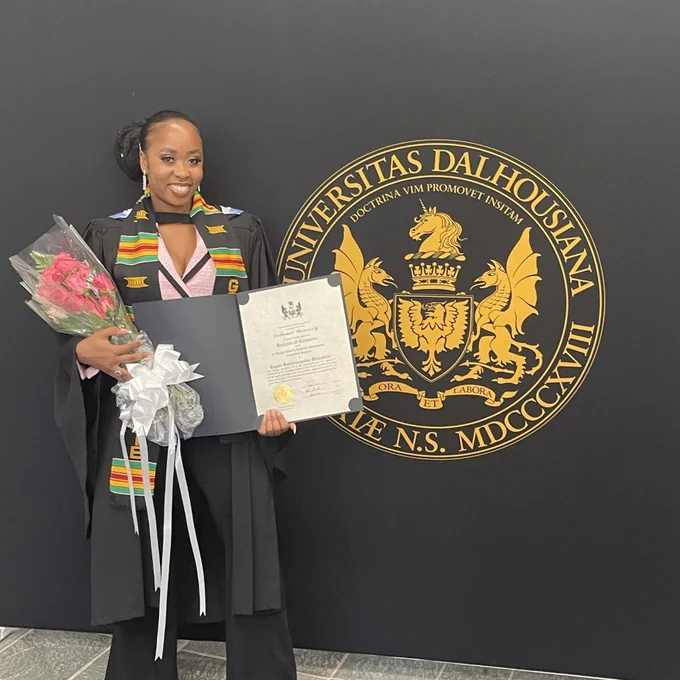 Kayler Mutyabule, a Black woman with her long hair pulled back, stands in front of a black wall with the Dalhousie crest on it. She is wearing a black graduation robe with a brightly-coloured kente sash overtop of a pink blazer and black pants with sparkly earrings and hair clips. She holds her diploma and a bouquet of pink flowers and she is smiling at the camera.