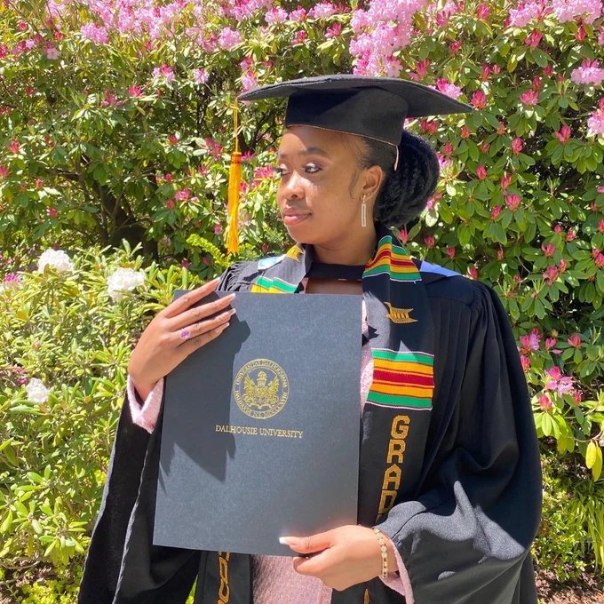 Kayler, a Black woman with her long hair pulled back, stands outdoors surrounded by greenery and pink flowers. She is wearing a black graduation robe and cap with a brightly-coloured kente sash overtop of a pink blazer with sparkly accessories. She holds her diploma and gazes offscreen with a stoic expression.
