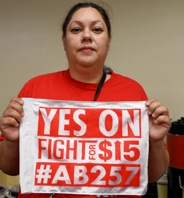 McDonald’s worker Crystal Orozco holding a sign that says YES ON AB 257 with the Fight for $15 logo. She's a middle aged latina who attended the last committee hearing after working 6pm-12am the night before the day long hearing. She's returned with workers from across the state. 