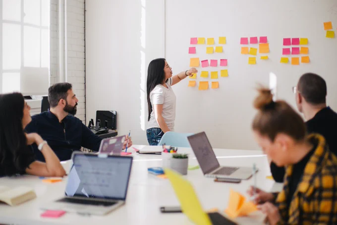 A group of people work around a table with laptops on it, looking at a woman who stands towards a white wall with colourful post-its on it.