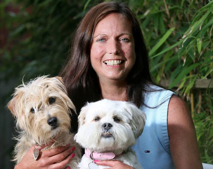 Close up of Nicola Whitehall, sat outside in a leafy area, holding two small dogs. 