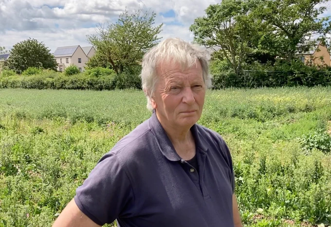 A man standing in a field. Trees and a hedgerow stand in the background. Beyond the trees are newly built houses.
