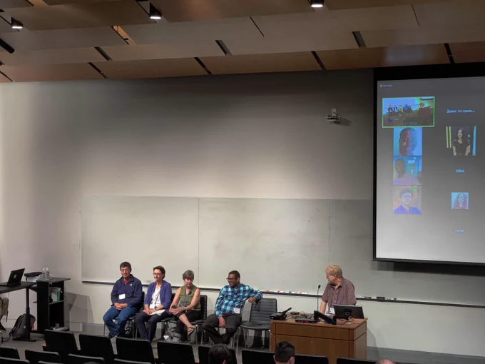 A room with one speaker standing at a table and four people sitting in nearby chairs. In the background the screen displays a portion of Zoom call.