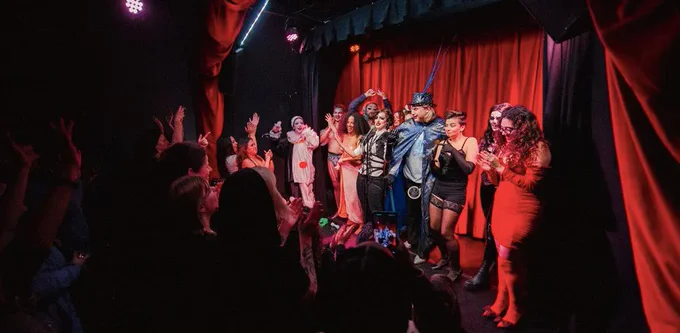 An audience applauding a group of cabaret artists in different costumes in front of a red curtain. 