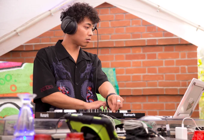 Image is of DJ ROMMEL. He is wearing headphones and is looking at a  laptop whilst altering one of the buttons on a mixing deck.