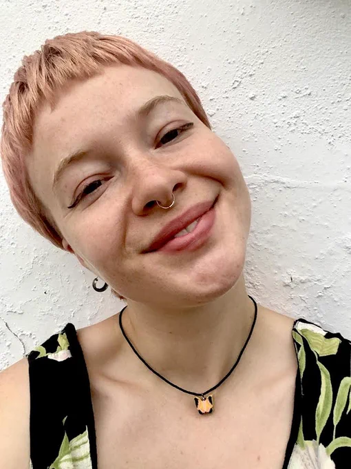 Image is a headshot of DJ So Faith. She has short rose pink hair, her head is tilted to the left, and she is smiling at the camera. 