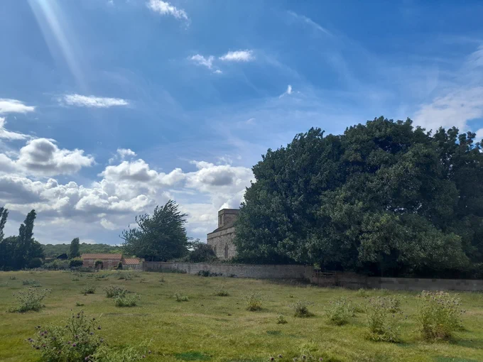 The photo depicts an overview of the old Owthorpe estate where Lucy Hutchinson and her husband Colonel John Hutchinson used to reside. It shows a view of the field where the Hall used to be, with a barn and Church in the background. 