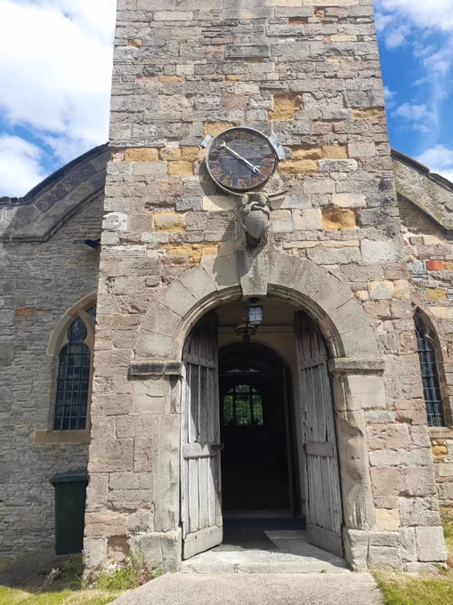The photo depicts the exterior of St. Margaret's Church at Owthorpe. The building appears to be made out of grey stone bricks and features a clock and sculpture over the entrance archway. 