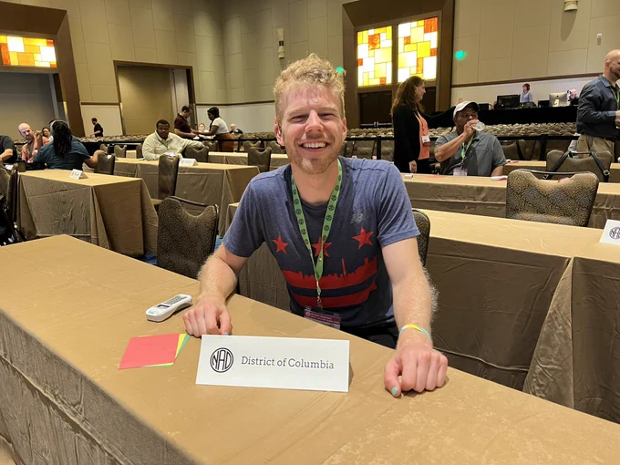 image description: Robb wearing DC flag shirt and sitting at table with "District of Columbia" placard