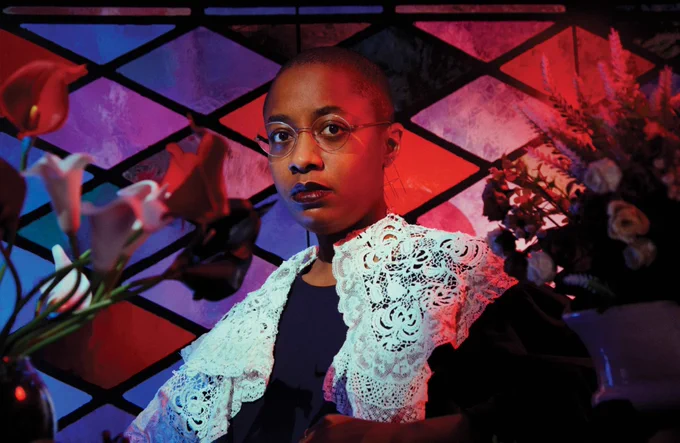 Cécile McLorin Salvant wearing a white lace shaw and glasses standing in front of a red, purple, and blue diamond background with flowers. Photo by Shawn Michael Jones.
