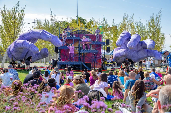 Squonk plays on their 3-tiered stage in the middle of the giant purple hand puppets on a field with people watching the performance. Photo courtesy of Squonk.