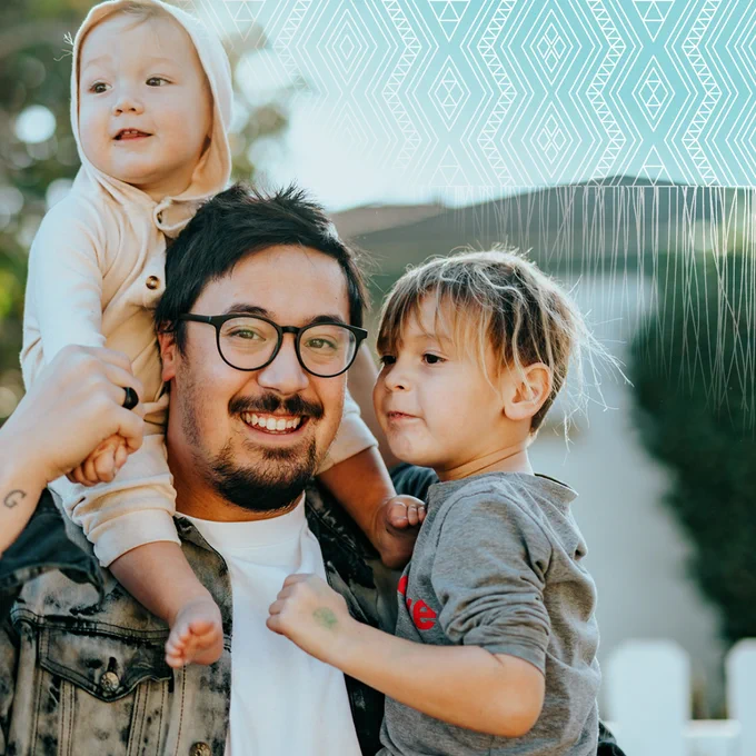 Image of three people looking at the camera. Two young children (tamariki) and one older male with glasses smiling looking the camera. One of the children is sitting on the male's shoulders. The other child is being carried by the male and looking away from the camera.