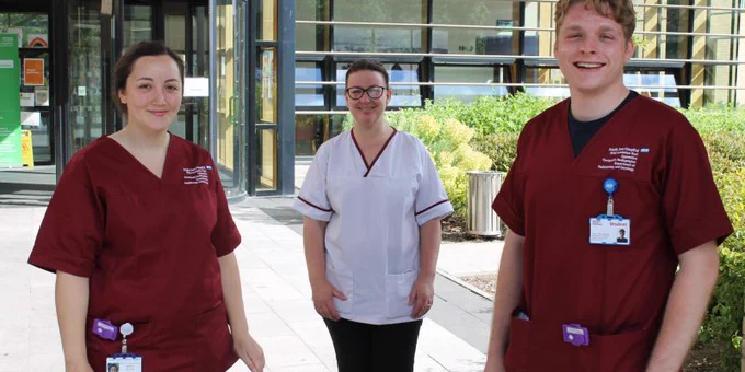 3 Healthcare workers stood outside the front of a hospital facing the camera. 