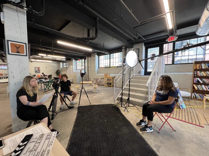 A woman is being interviewed on camera by another woman and a man is behind the camera.
