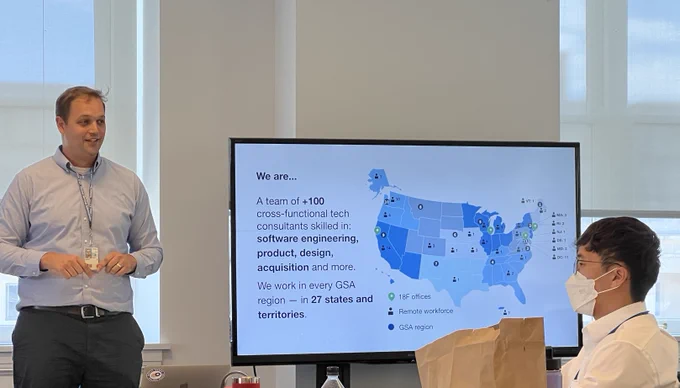  Joe Krzystan talks with the Fellows about 18F. The screen next to him shows a map of the US where 18F staff is located A team of  100 cross-functional tech consultants who work in 27 states and territories.