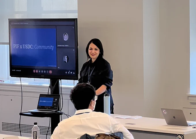 Rebeca Lamadrid stands next to a screen that shows a blue slide with the title "PIFxUSDC: Community". She smiles as she talks with the Fellows about the PIF program.