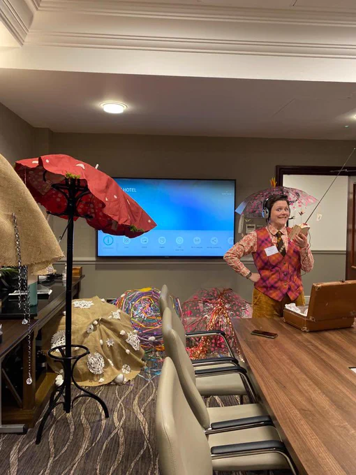 Gemma in a bright waistcoat, holding a box like a radio in front of a vintage suitcase, with decorated umbrellas around the edge of the room.