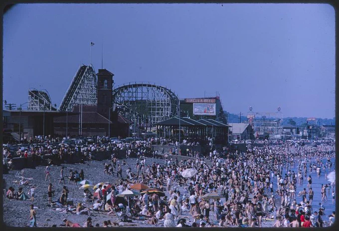 Photograph of a crowd of people on Revere Beach, with the Cyclone rollercoaster in the background. 