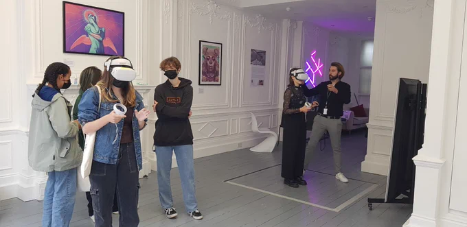 A room full of people, two of whom are wearing VR headsets. The room is white and grand-looking, and has colourful artwork hung on the walls, and LED lighting.