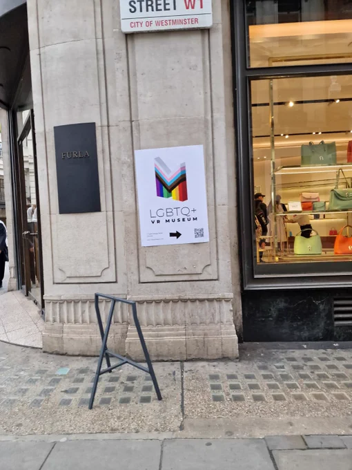 A poster with the words "LGBTQ  VR museum" and a colourful M-shaped logo, affixed to a stone wall on a London street 
