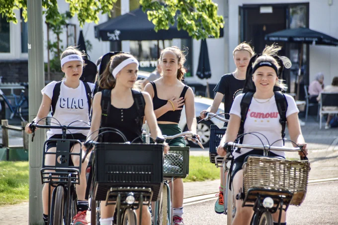 A group of five teenage girls cycle together in a Dutch city. They are dressed for some kind of sporting practice.