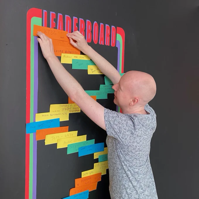 A photograph of gallery visitor Mavit placing his winning score at the top our leaderboard, which is a painted area on the gallery wall. All the names of recent visitors and their scores have been added on brightly coloured pieces of paper. His current personal best score is 12877. Outstanding!