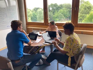 Three people working at a desk in an office, the window looks out onto trees. 