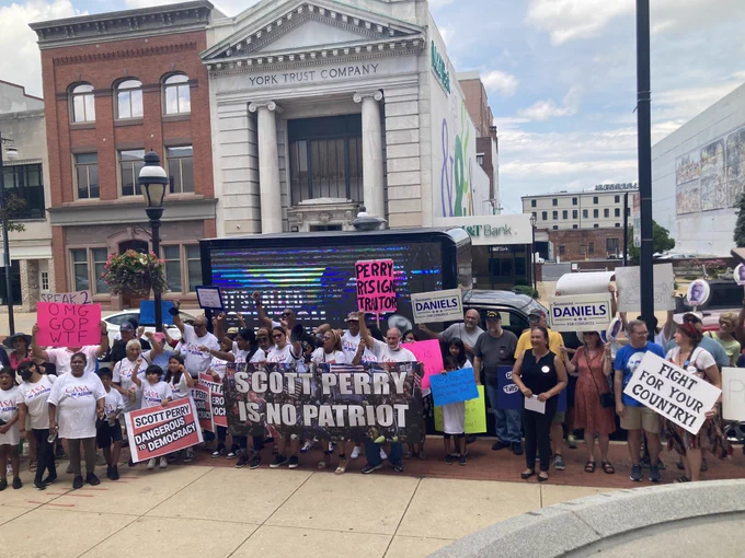 Large crowd of people including Shamaine gathered around a banner reading SCOTT PERRY IS NO PATRIOT